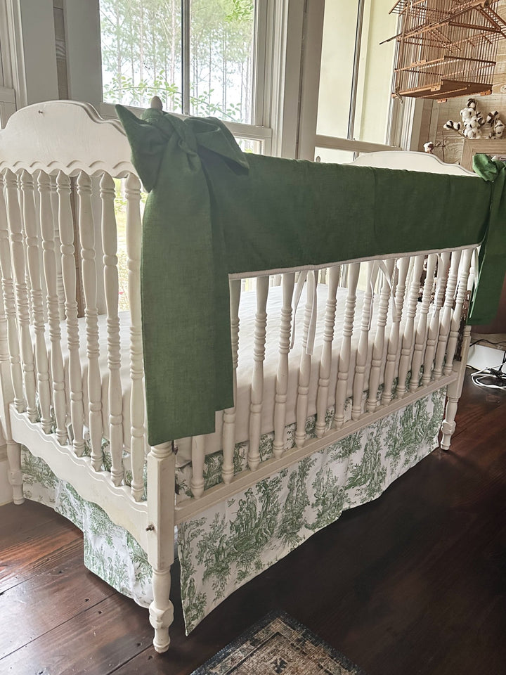 Angled view of the solid green tailored rail cover with a long green bow sash draped over the crib rail
