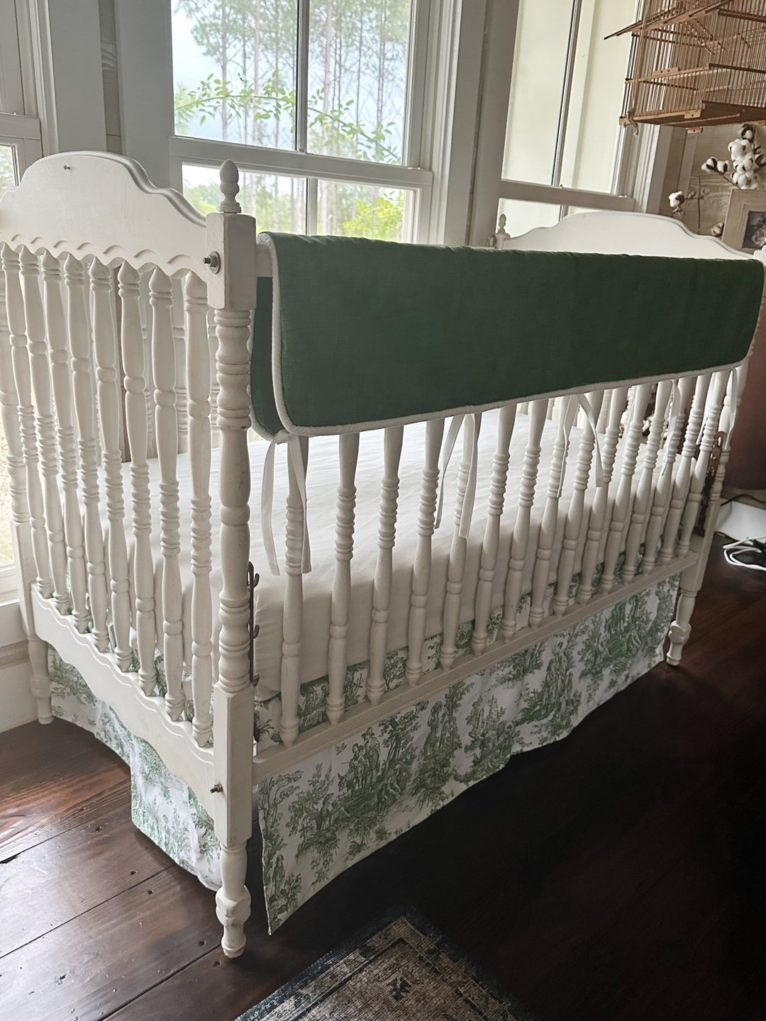 Angled view of the cypress green tailored rail cover and green toile crib skirt on a white spindle crib