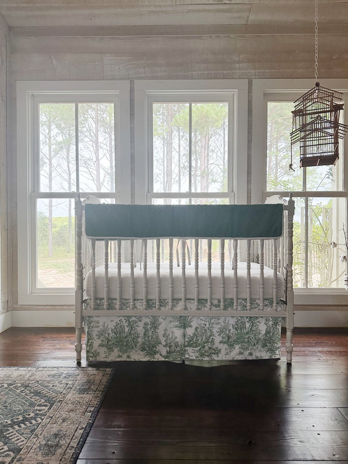Green toile and cypress green tailored crib bedding styled in a farmhouse nursery with pine trees beyond the windows