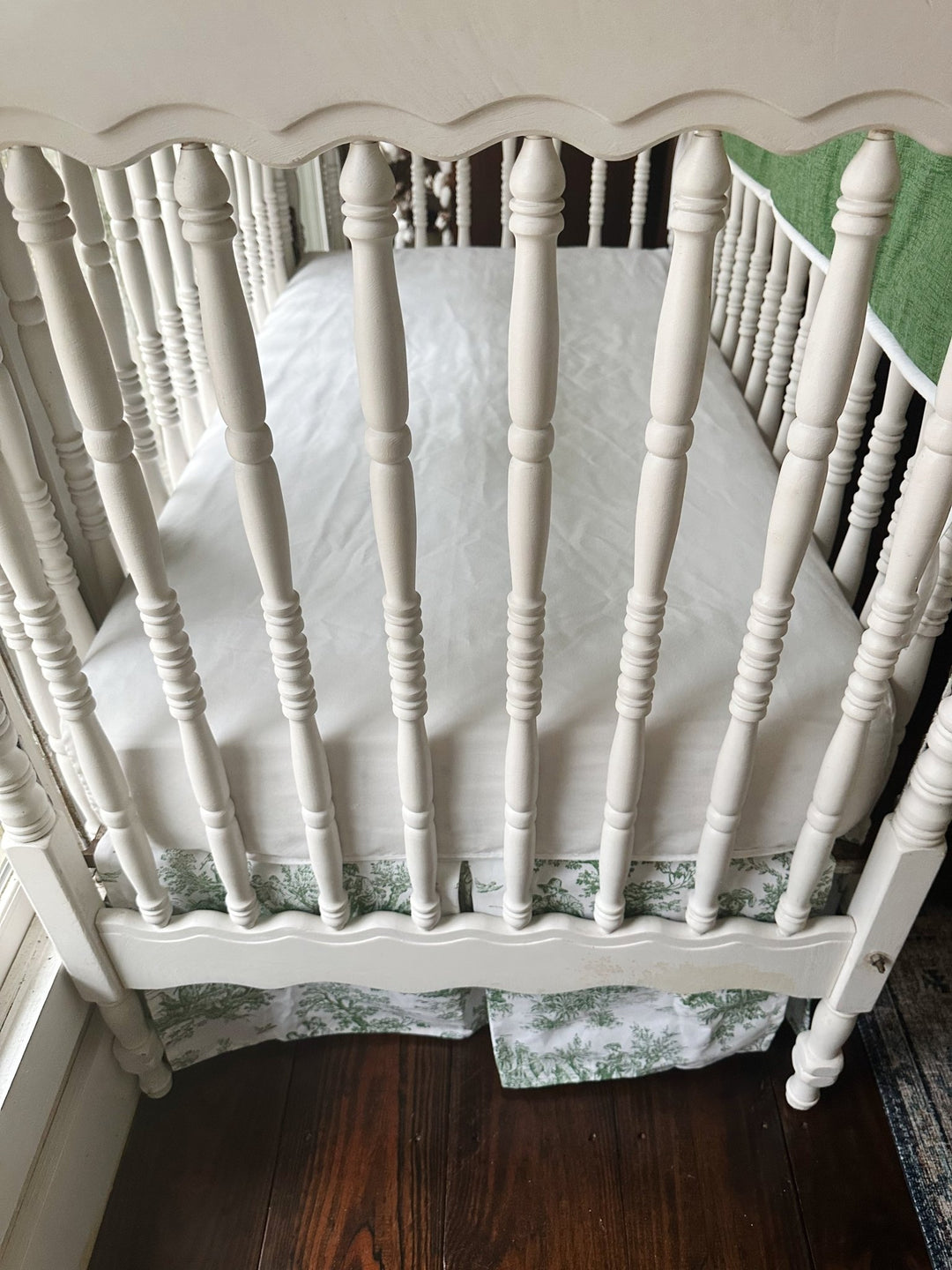 Overhead view of a white fitted crib sheet with the green toile tailored crib skirt below