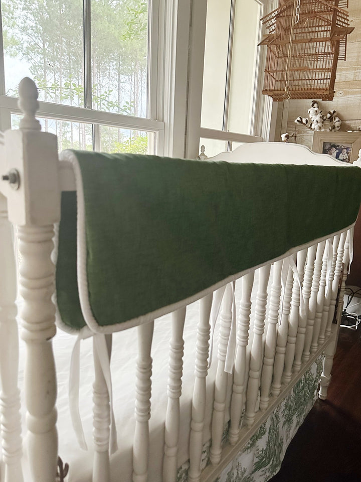 Cypress green tailored crib rail cover with white cording draped along the top rail of a white spindle crib