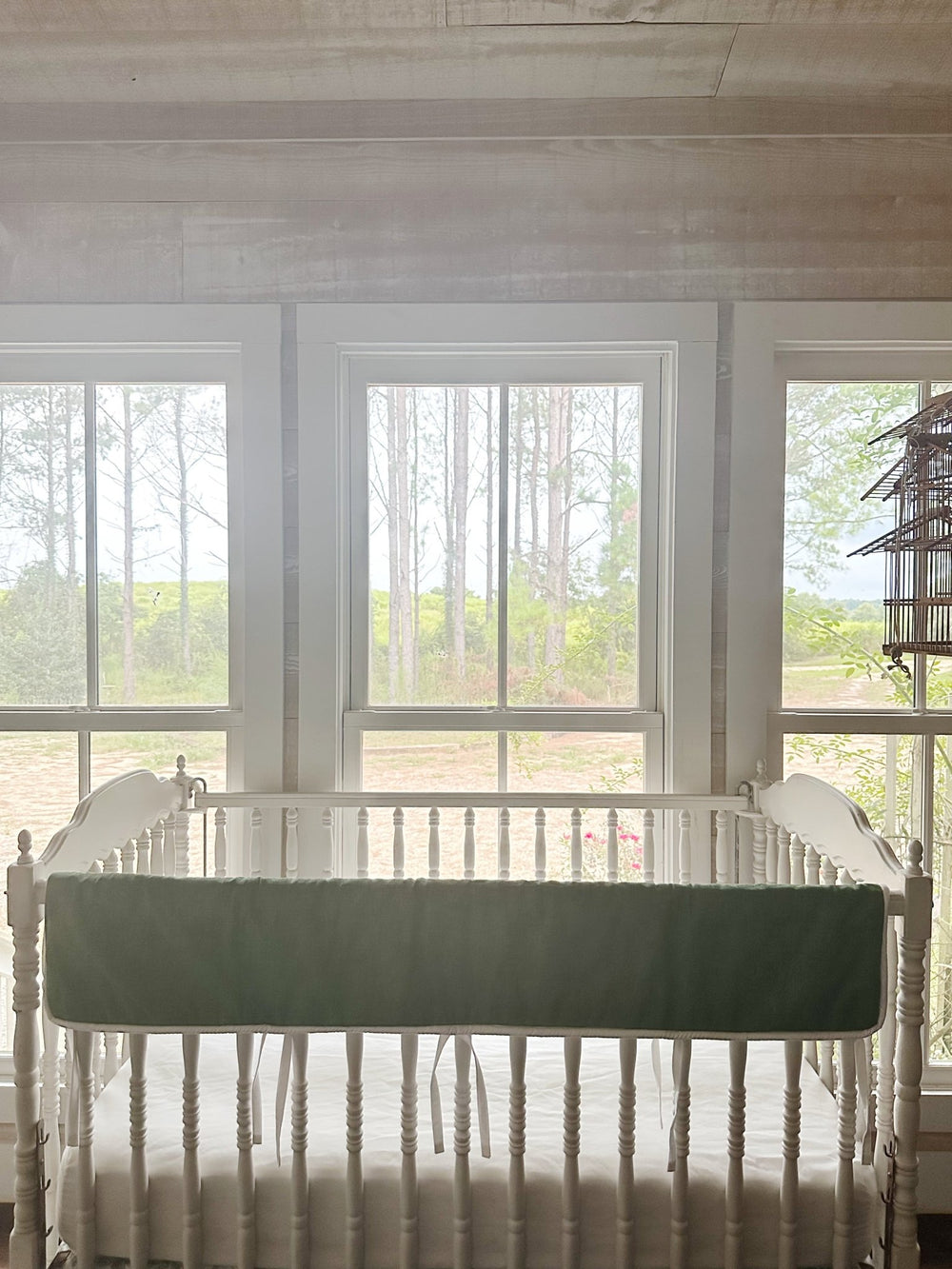 Cypress green tailored crib rail cover on a Jenny Lind style crib in a bright nursery with pine trees beyond the windows