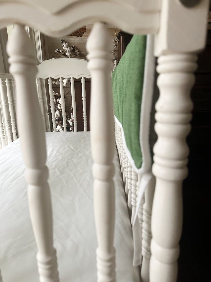 Cypress green crib rail cover seen through white crib spindles with cotton stems in the background