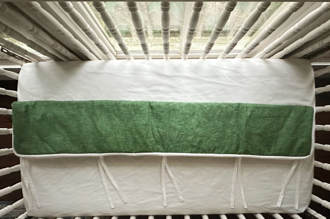 Overhead view of the cypress green tailored crib rail cover laid flat across a white fitted crib sheet