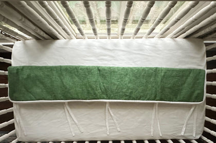 Overhead view of the cypress green tailored crib rail cover laid flat across a white fitted crib sheet