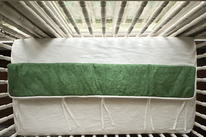 Overhead view of the cypress green tailored crib rail cover laid flat across a white fitted crib sheet