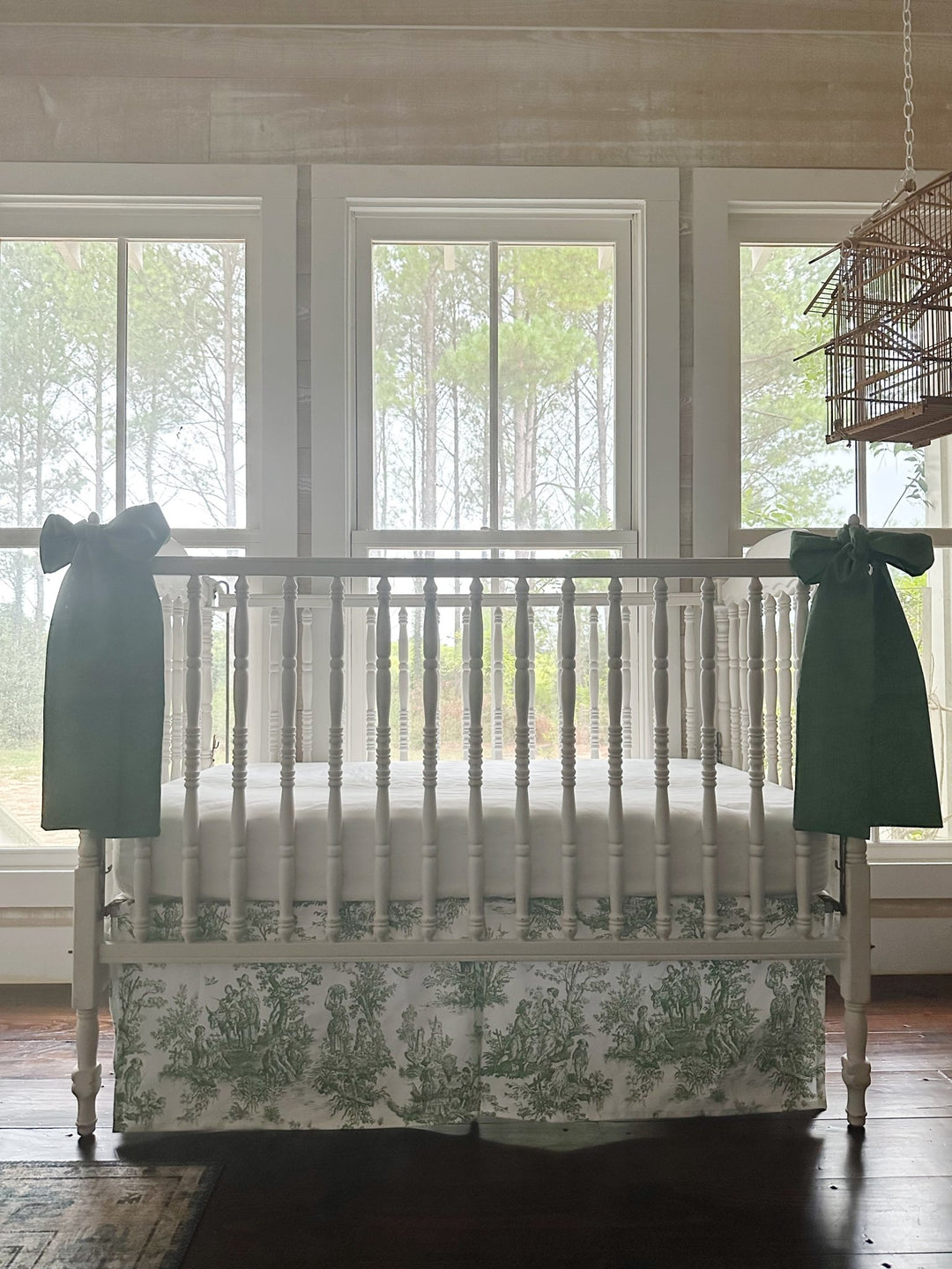 White crib dressed with a green toile box-pleat skirt and a large cypress green bow on each front post