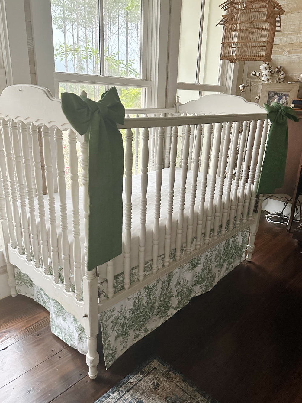 Green toile tailored crib skirt and two cypress green bows on a white spindle crib, seen from the corner