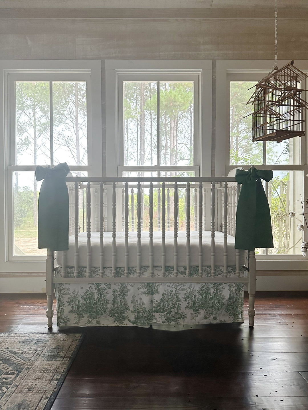 Green toile crib skirt and cypress green bows in a farmhouse nursery with pine woods beyond the windows
