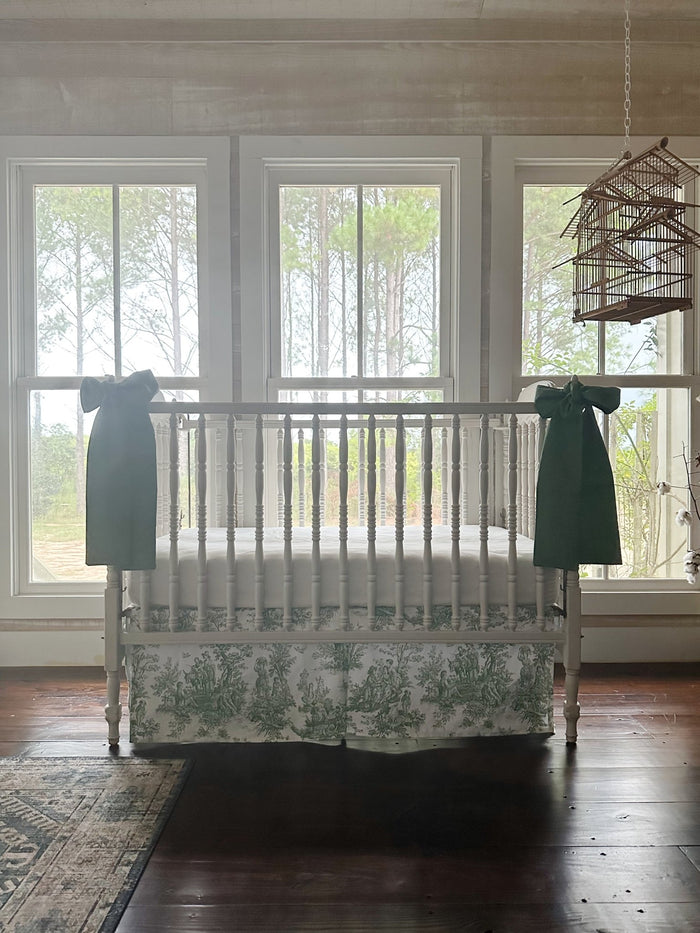 Green toile crib skirt and cypress green bows in a farmhouse nursery with pine woods beyond the windows