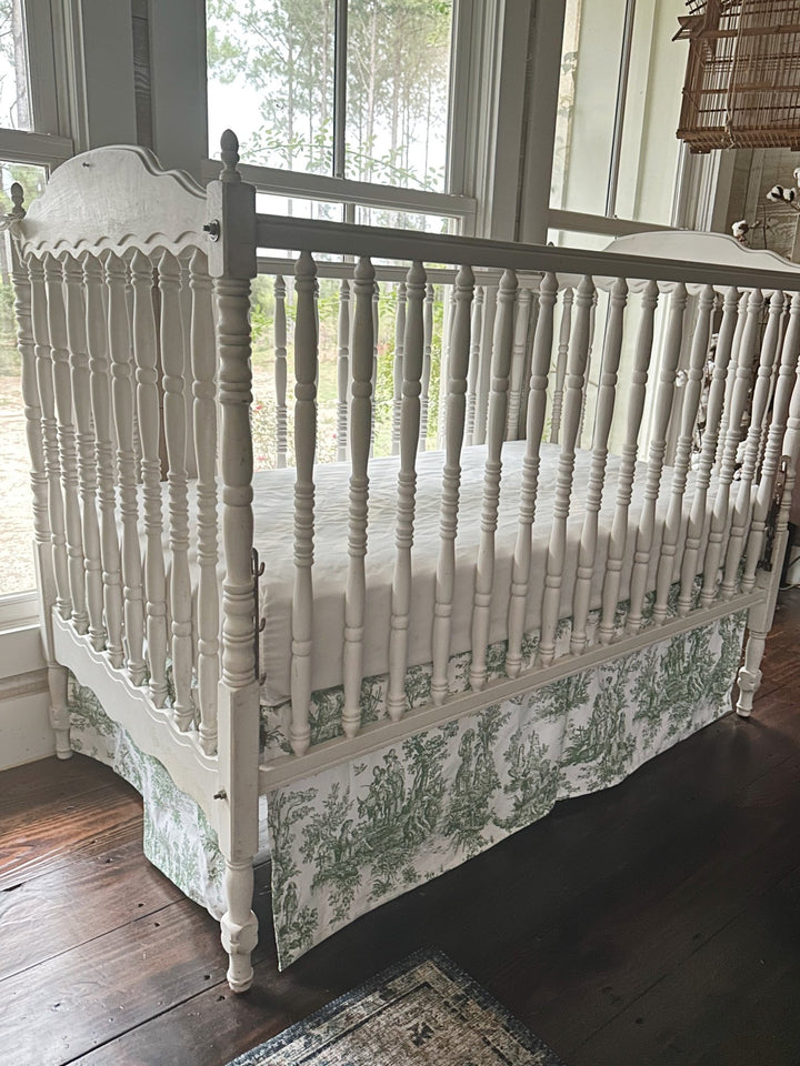 Green toile tailored crib skirt on a white spindle crib, seen from the corner with the pleated panels falling straight