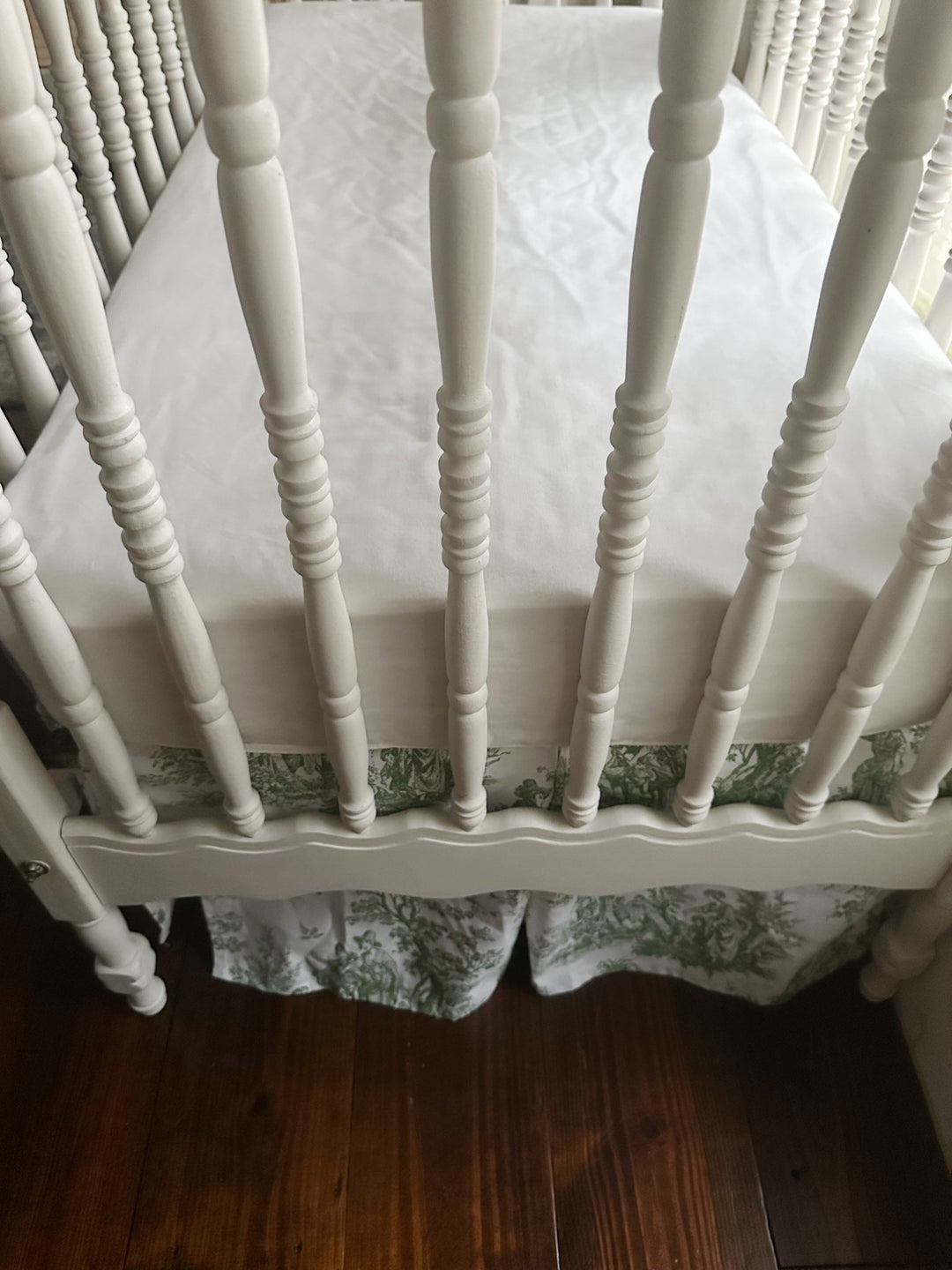 Looking down into the crib at the white sheet with the toile skirt deck below the mattress platform