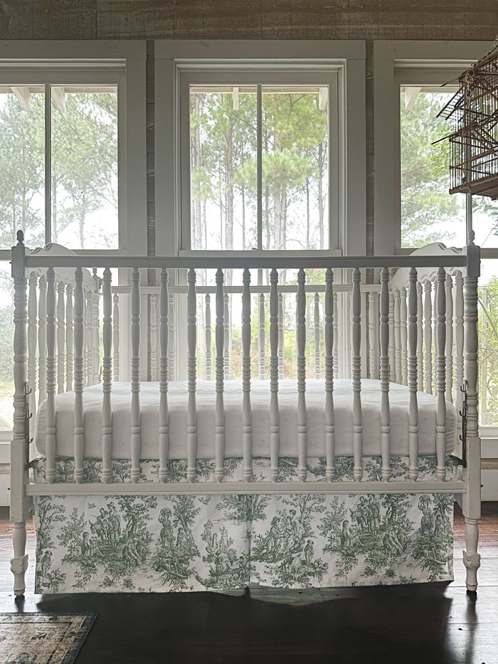 Green toile crib skirt seen straight on beneath a white fitted crib sheet