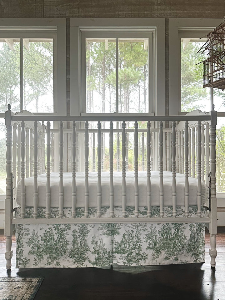 Green toile crib skirt seen straight on beneath a white fitted crib sheet