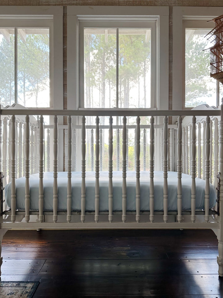 Front view of a white spindle crib made up with the Heirloom Blue fitted crib sheet against tall nursery windows - High Cotton Textile