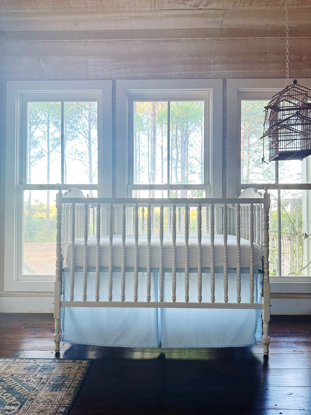 Farmhouse nursery with three tall windows looking out on the pines and a white crib dressed in a heirloom blue tailored crib skirt - High Cotton Textile