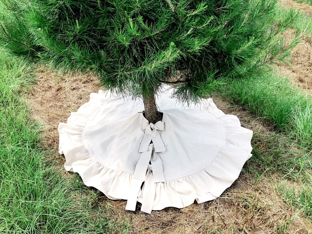 Natural osnaburg ruffled Christmas tree skirt spread around a pine trunk, cotton ties knotted into bows at the front opening