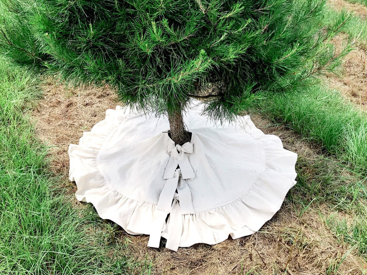 Natural osnaburg ruffled Christmas tree skirt spread around a pine trunk, cotton ties knotted into bows at the front opening