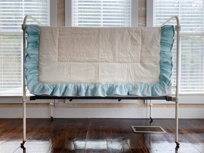 Ivory ruffled baby quilt with a gathered mist blue ruffle draped over an antique iron crib rail