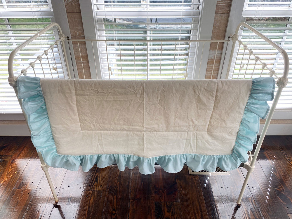 Ivory and mist ruffled crib quilt hung across the front rail of a white iron crib