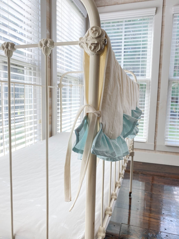 Ivory cotton tie knotted at the corner post of an iron crib beside the mist blue ruffle