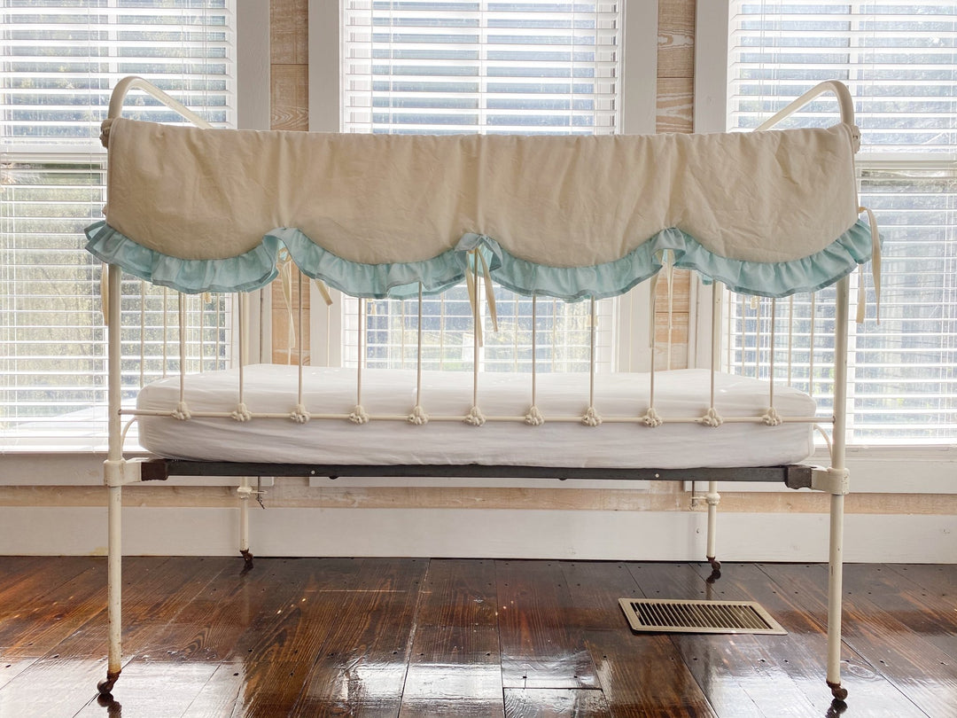 Ivory scalloped crib rail cover with a soft mist blue ruffle on an antique white iron crib