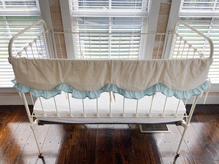 Ivory and mist scalloped crib rail cover shown across the long front rail of a vintage iron crib