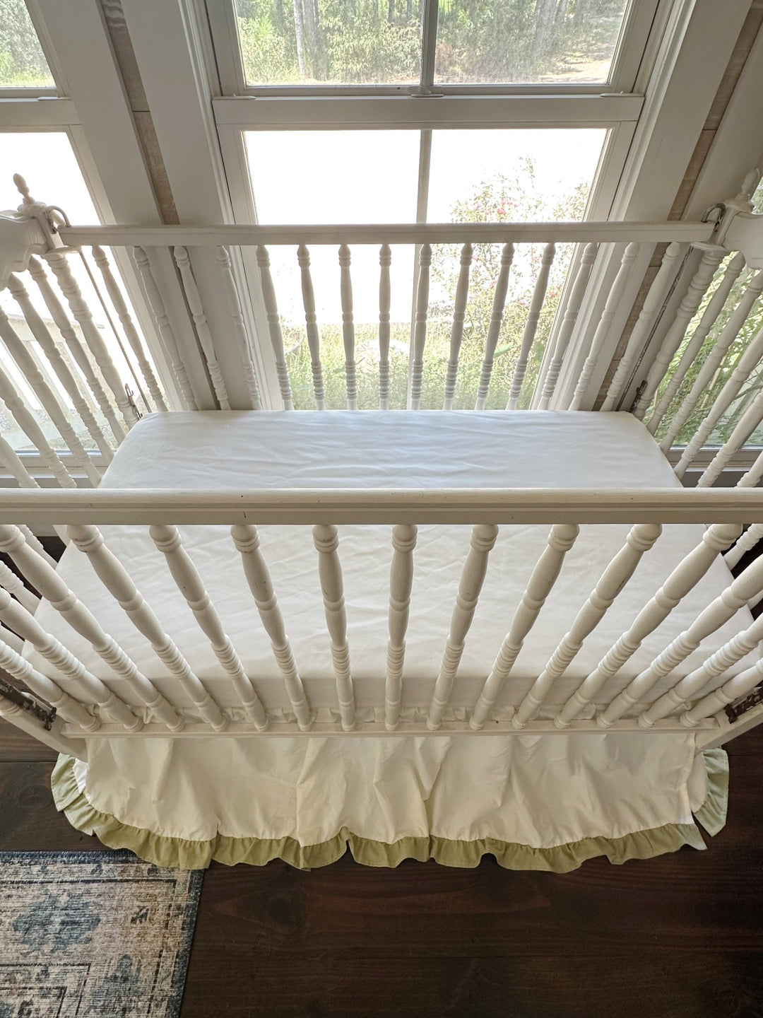 Overhead view of an ivory ruffled crib skirt with sage green trim in a sunlit gender neutral nursery