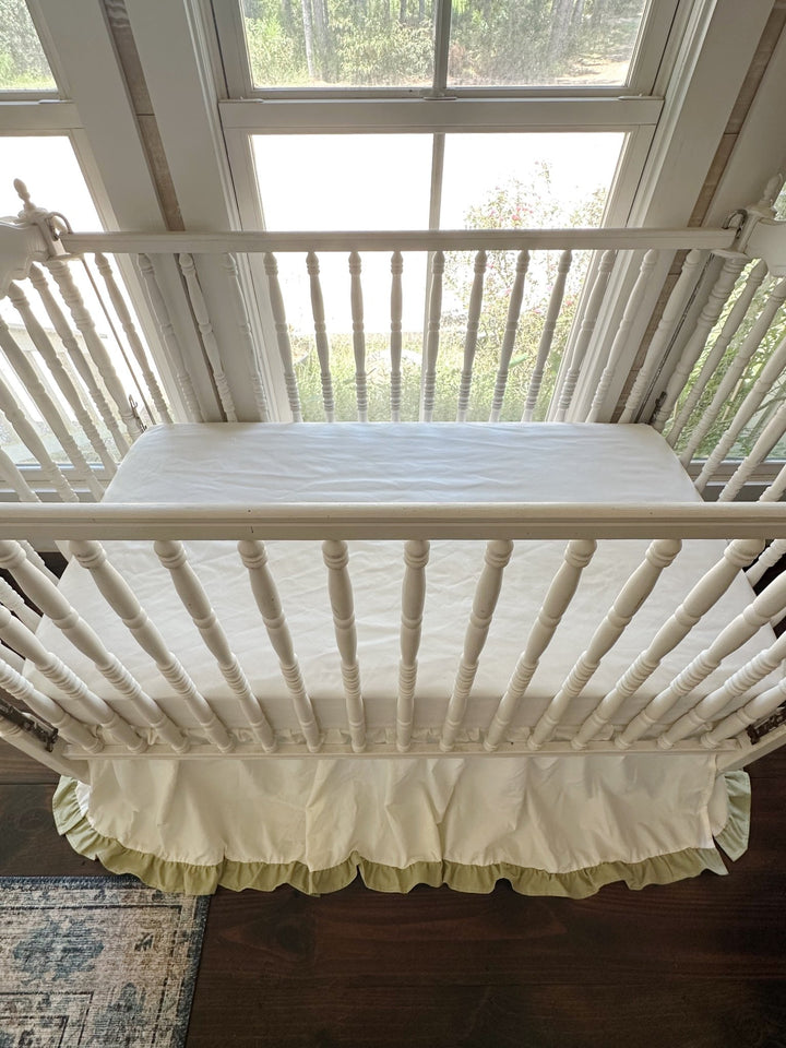 Overhead view of an ivory ruffled crib skirt with sage green trim in a sunlit gender neutral nursery