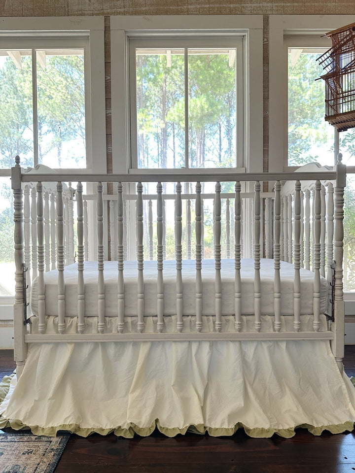 Close front view of an ivory crib skirt with sage green ruffle trim on an antique white spindle crib