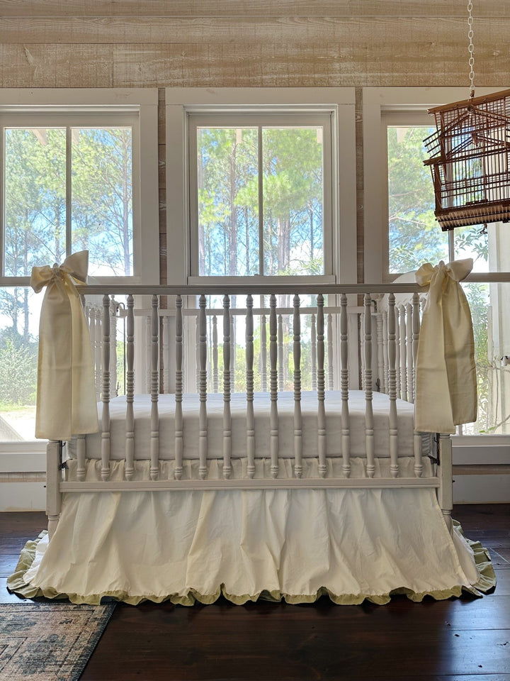 Front view of a neutral crib bedding set with ivory ruffled crib skirt, sage green hem and two ivory bow sashes