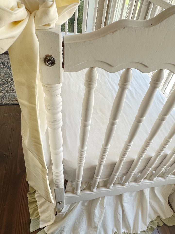 Ivory bow sash draped over the crib rail with the gathered ivory and sage crib skirt below