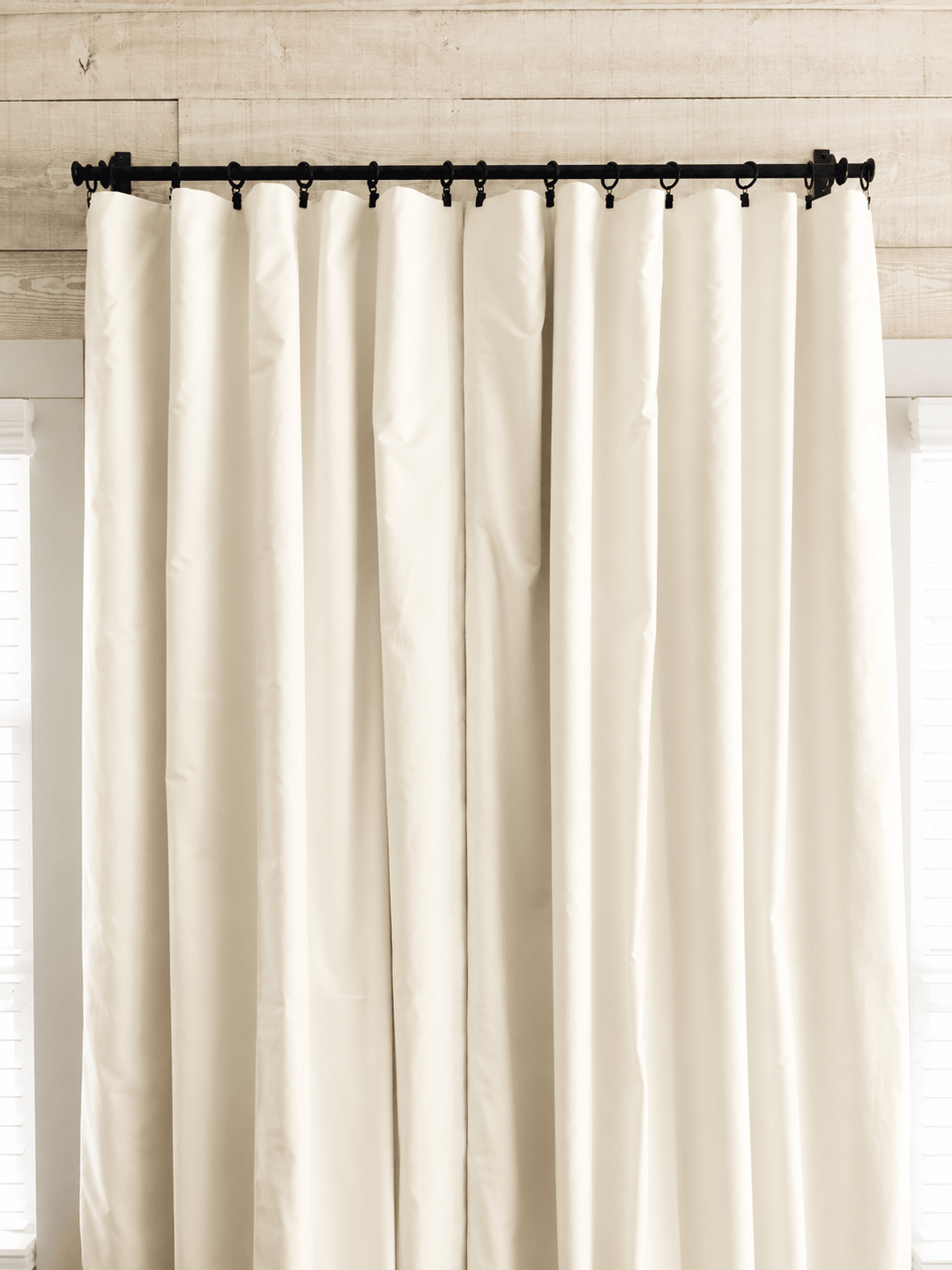 Ivory blackout curtain panels drawn closed on a black iron rod with clip rings, against a whitewashed plank wall