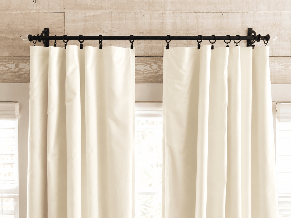 Ivory panels parted on a black rod with rings, daylight and greenery showing through the window between them