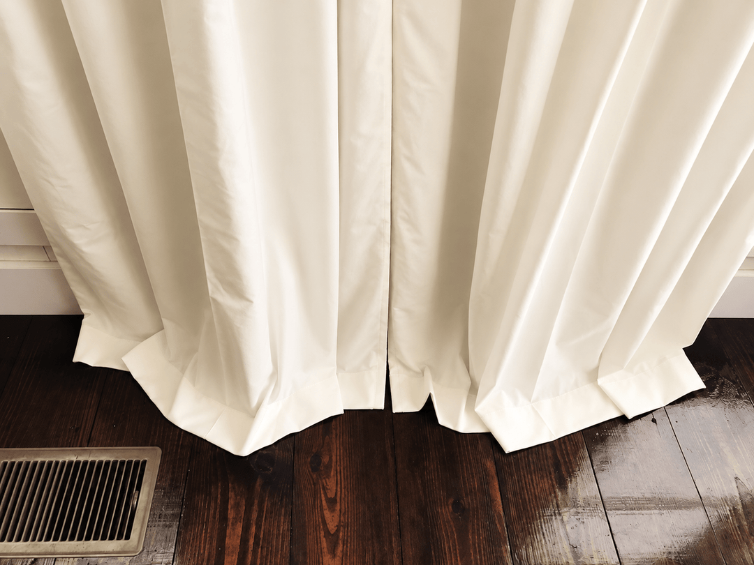Hem detail of ivory blackout curtains breaking against a dark stained wood floor beside a floor vent