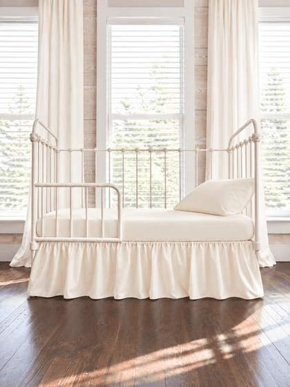 Ivory farmhouse toddler bed skirt gathered on all four sides of a cream iron crib converted to a toddler bed, with matching ivory sheet and pillow, ivory curtains behind - High Cotton Textile