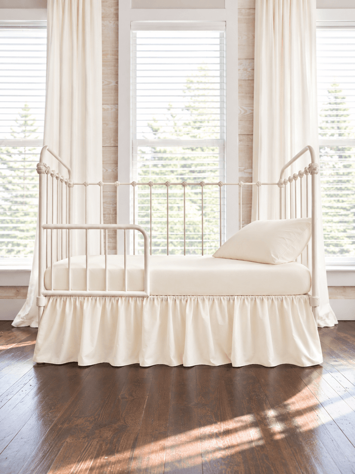 Ivory farmhouse toddler bed skirt gathered on all four sides of a cream iron crib converted to a toddler bed, with matching ivory sheet and pillow, ivory curtains behind - High Cotton Textile
