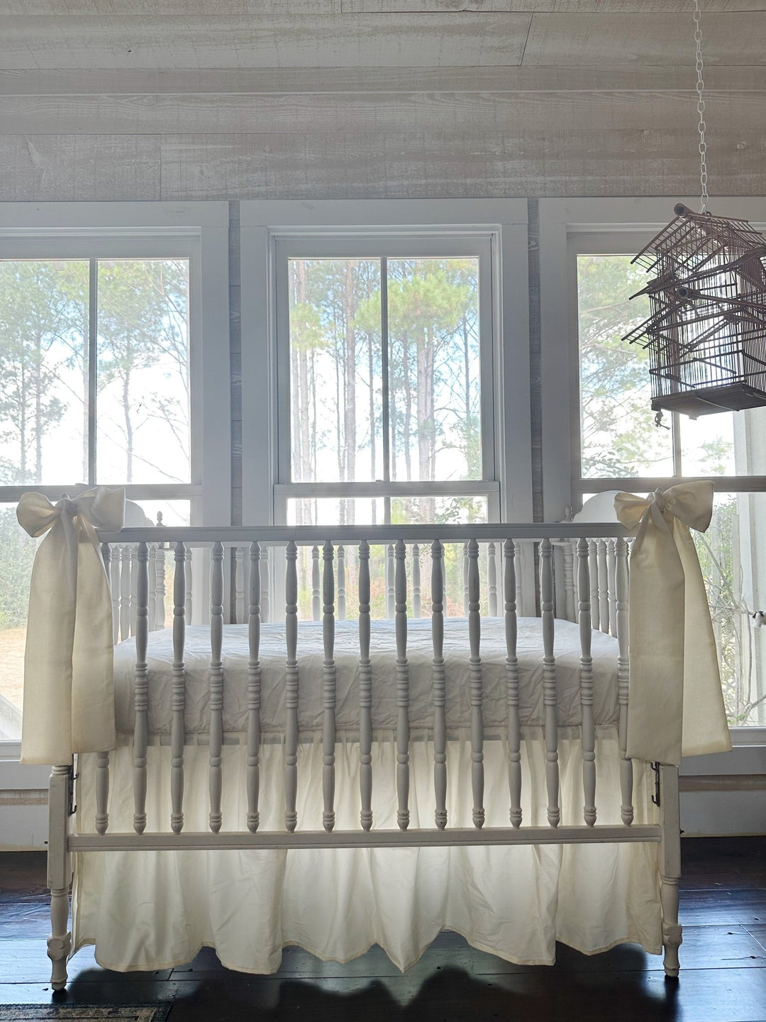 Ivory farmhouse crib skirt and two ivory bows on a white spindle crib, straight-on in front of three tall windows