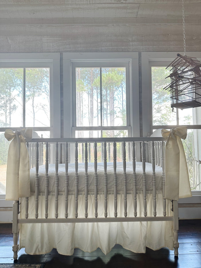 Ivory farmhouse crib skirt and two ivory bows on a white spindle crib, straight-on in front of three tall windows