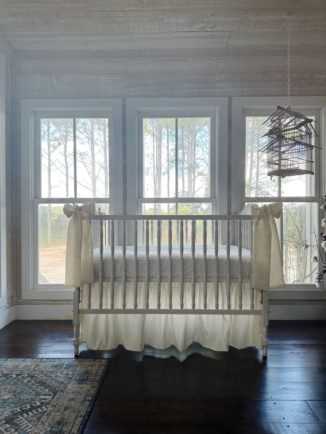 Nursery with whitewashed plank walls and a hanging birdcage, white spindle crib dressed in a gathered ivory skirt with ivory bows at both corners