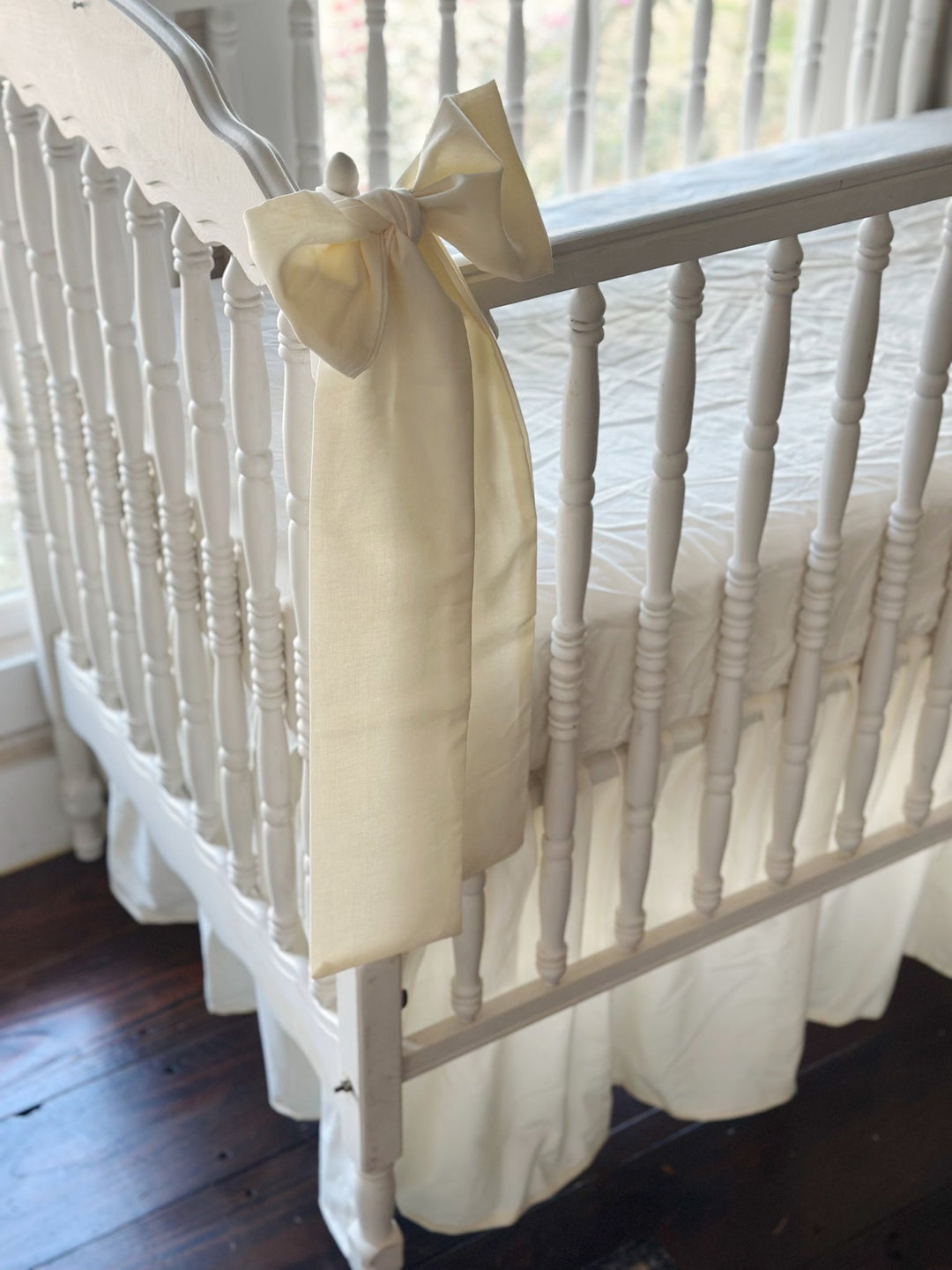 Close-up of an ivory cotton bow knotted at the crib's corner finial, long sash falling over the gathered ivory skirt