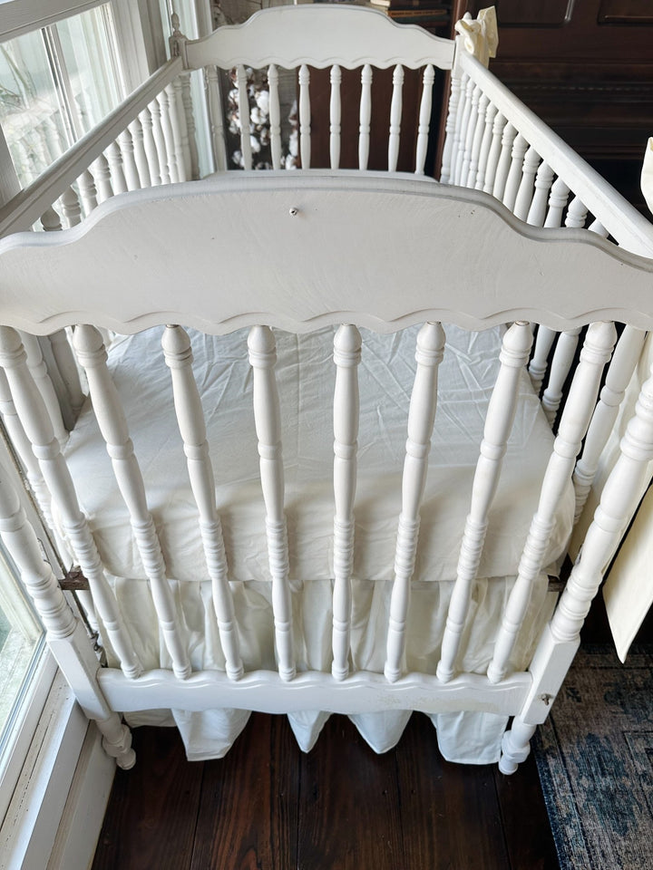 Looking down over the scalloped end of the crib at an ivory fitted mattress with the gathered ivory skirt below