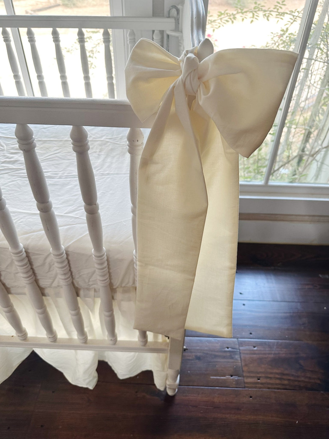 Ivory bow with a long sash tied at the crib post beside a sunny window, farmhouse skirt gathered to the wood floor