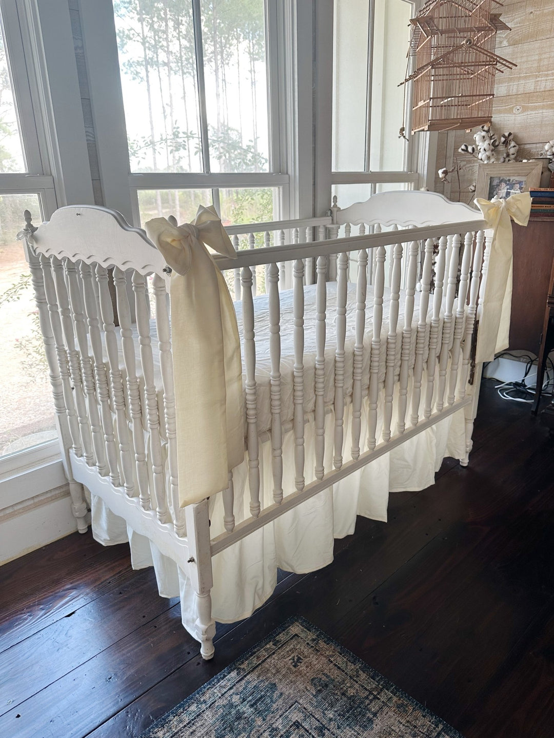 Three-quarter view of the ivory skirt and bow set on a white spindle crib by the windows, bow tails hanging down the corner posts