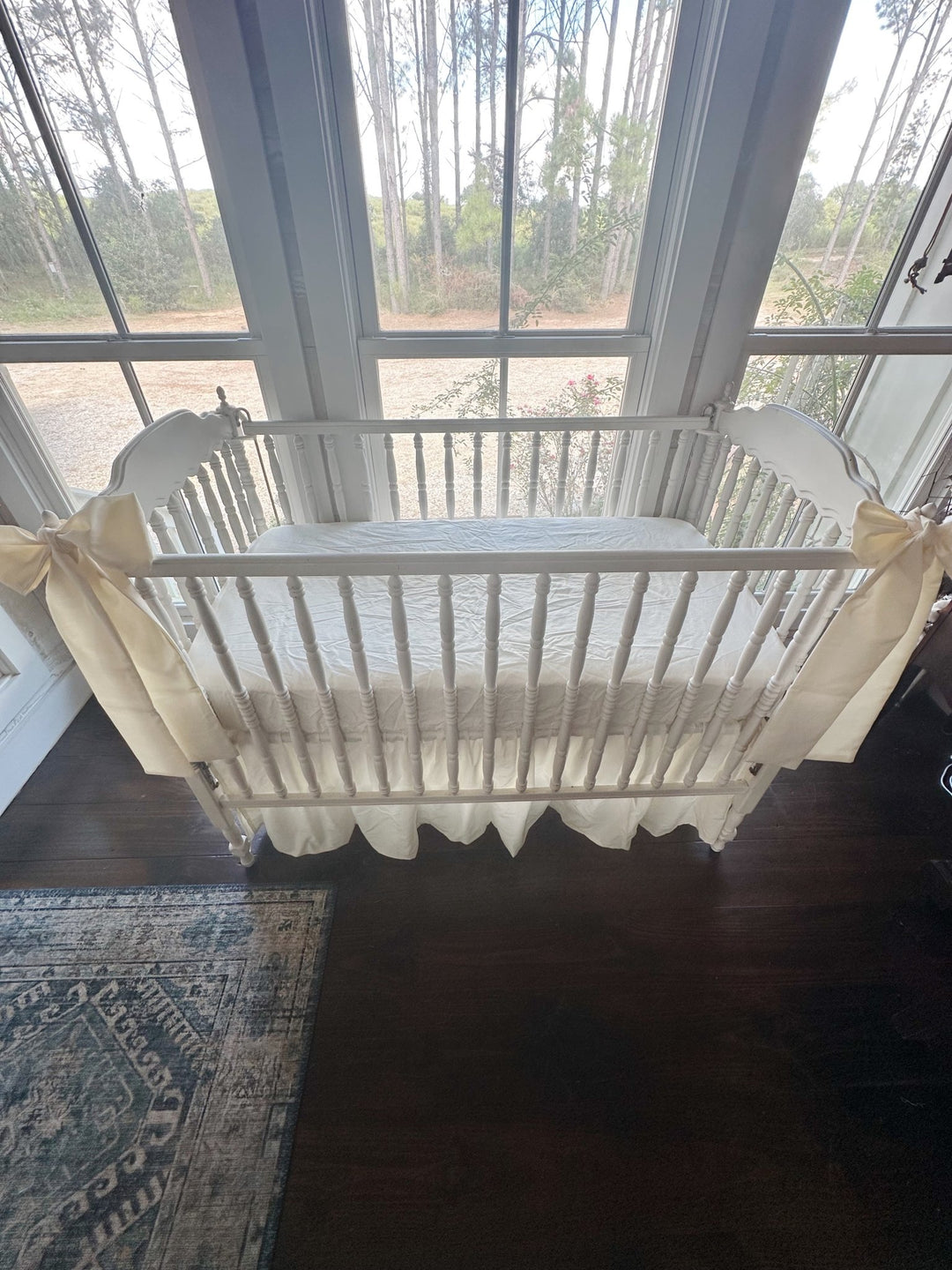 Backlit view from above of the ivory skirt and corner bows on a white spindle crib in front of three windows