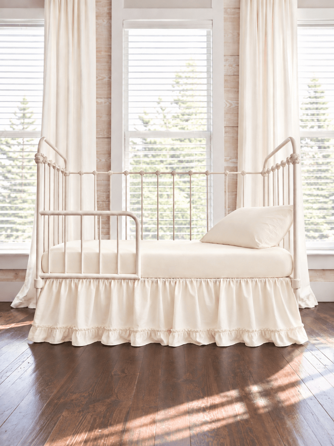 Ivory ruffled toddler bed skirt with a gathered body and ruffled hem on a cream iron crib converted to a toddler bed, ivory sheet, pillow and curtains - High Cotton Textile