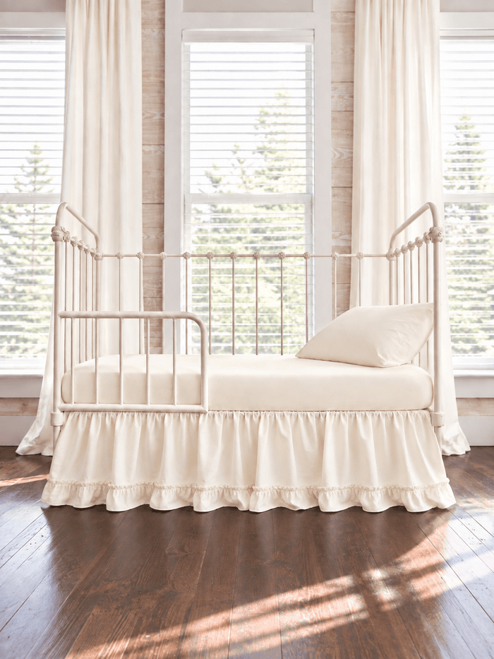 Ivory ruffled toddler bed skirt with a gathered body and ruffled hem on a cream iron crib converted to a toddler bed, ivory sheet, pillow and curtains - High Cotton Textile