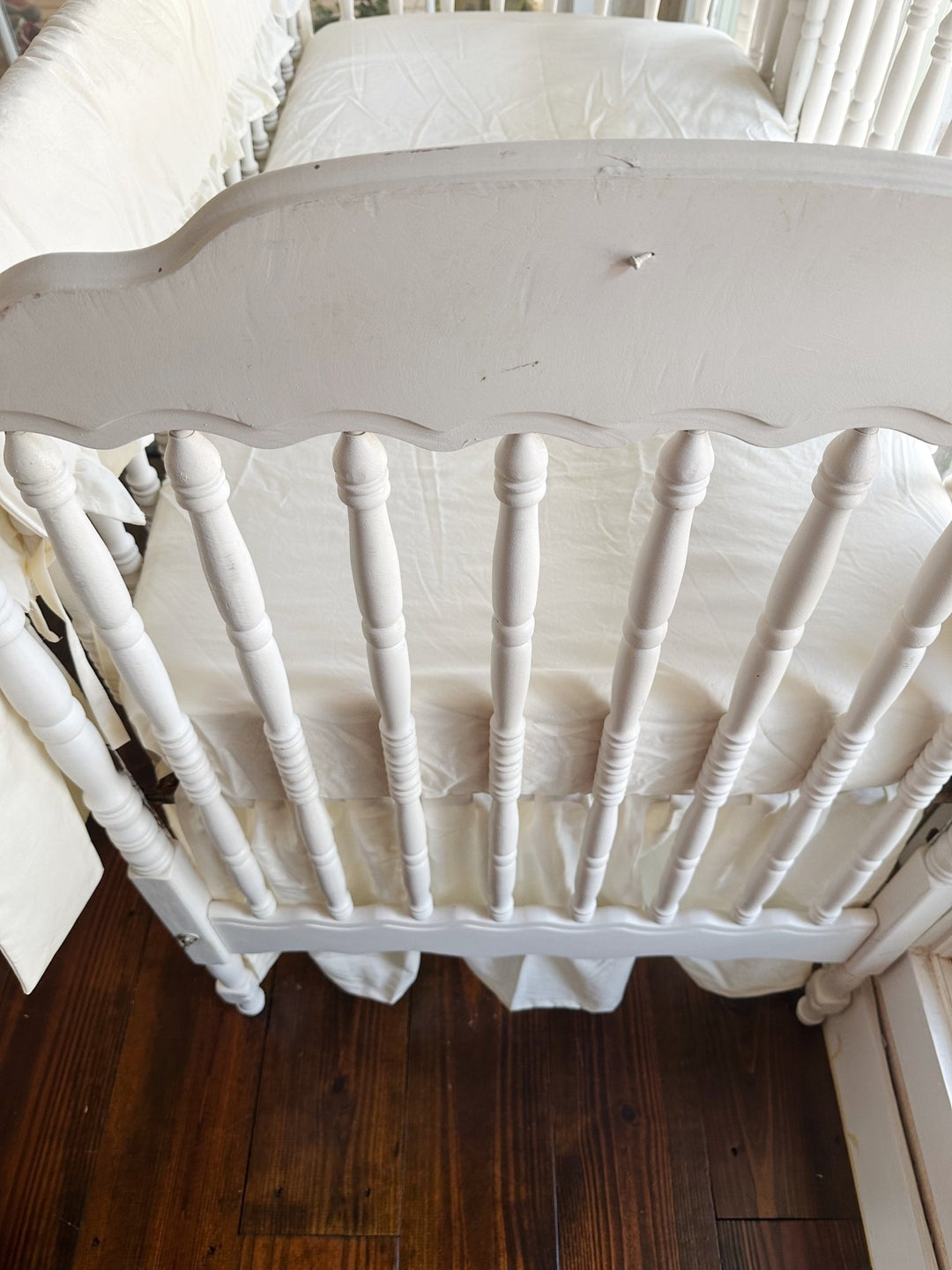 Looking down over the scalloped end of the crib at an ivory fitted mattress, gathered ivory skirt and the rail cover along the side