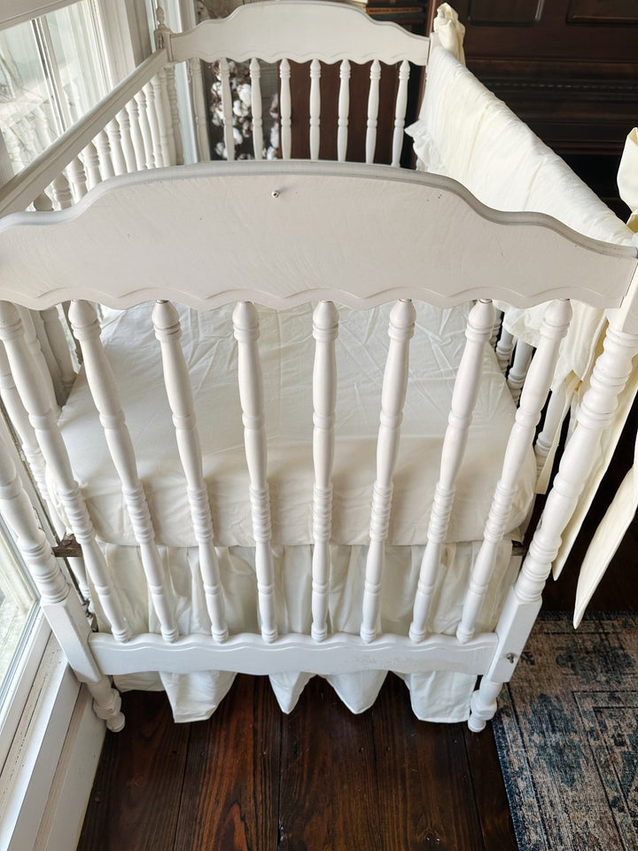 Close overhead view of the scalloped crib headboard with ivory skirt gathered below the mattress and the rail cover at the edge