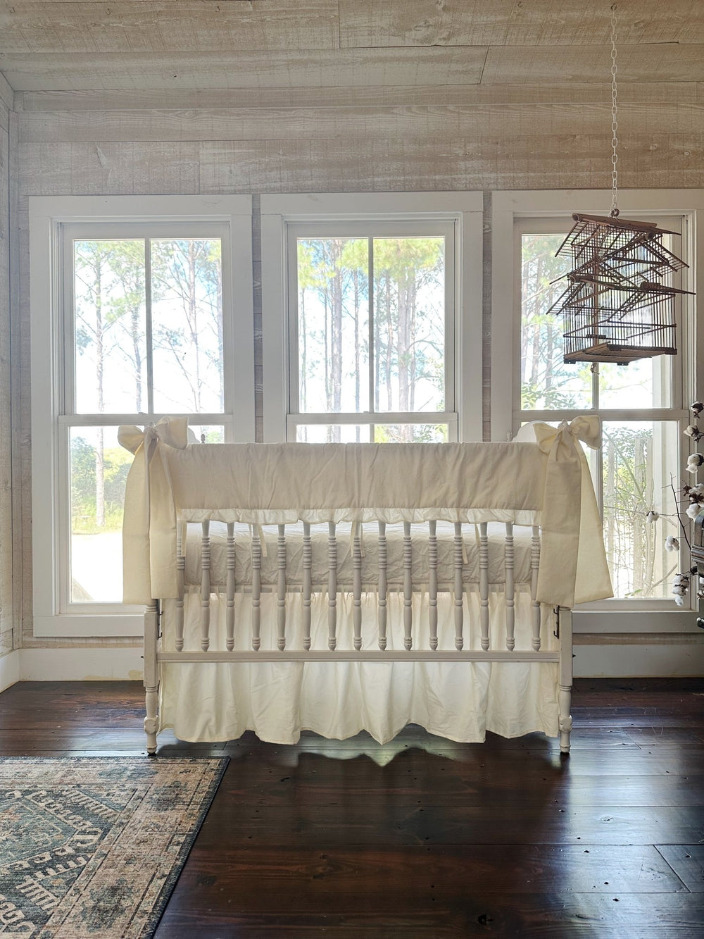 Nursery with whitewashed plank walls and a hanging birdcage, white spindle crib dressed in ivory ruffled rail cover, gathered skirt and corner bows